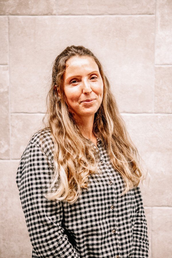 Eine Frau mit langem, lockigem, blondem Haar trägt ein schwarz-weiß kariertes Hemd vor einer Steinswand.