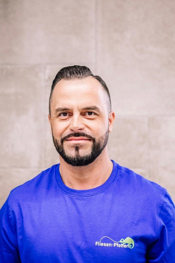 Ein Mann mit dunklem Haar, Bart und Schnurrbart trägt ein blaues T-Shirt mit einem Logo und allem auf einem neutralen Hintergrund.