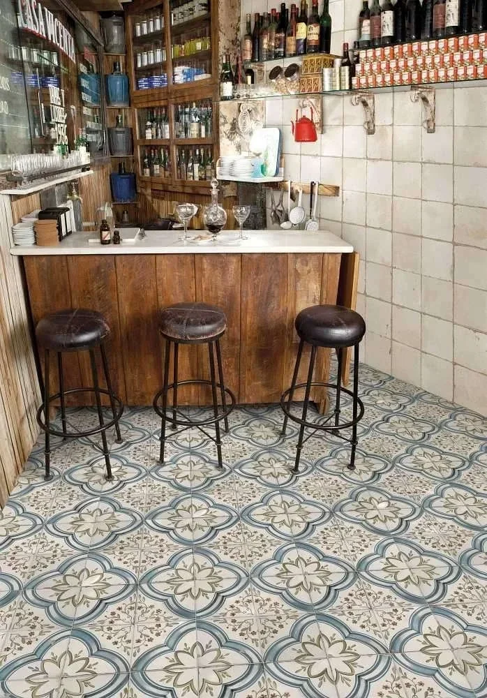 Ein Tresenbereich in einer Bar mit drei Barhockern, einem Holzregal mit Flaschen und Gläsern, sowie einer Wand mit Flaschen, Dosen und Geschirr.