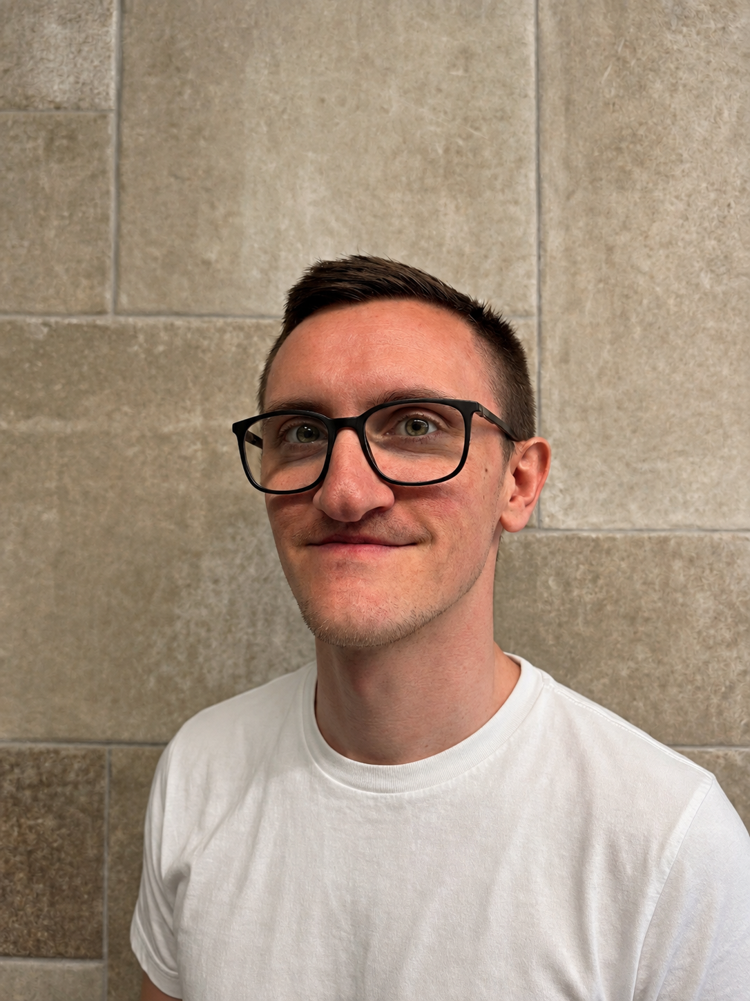 Porträt eines Mannes mit Brille, kurzhaarig, in einem weißen T-Shirt vor einer Steimauer.