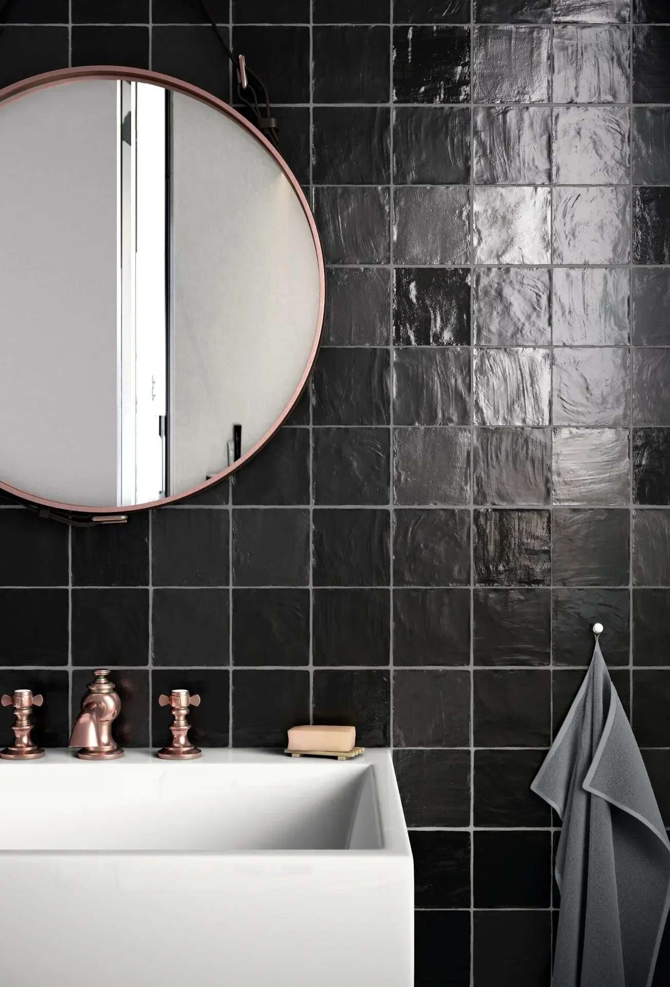 Detailaufnahme eines Badezimmerspiegels und eines Waschbeckens mit Armaturen aus roségold, schwarze Wandfliesen, eine weiße Handtuchhalterung mit grauem Handtuch und einem Seifenstück auf einem Waschtisch.