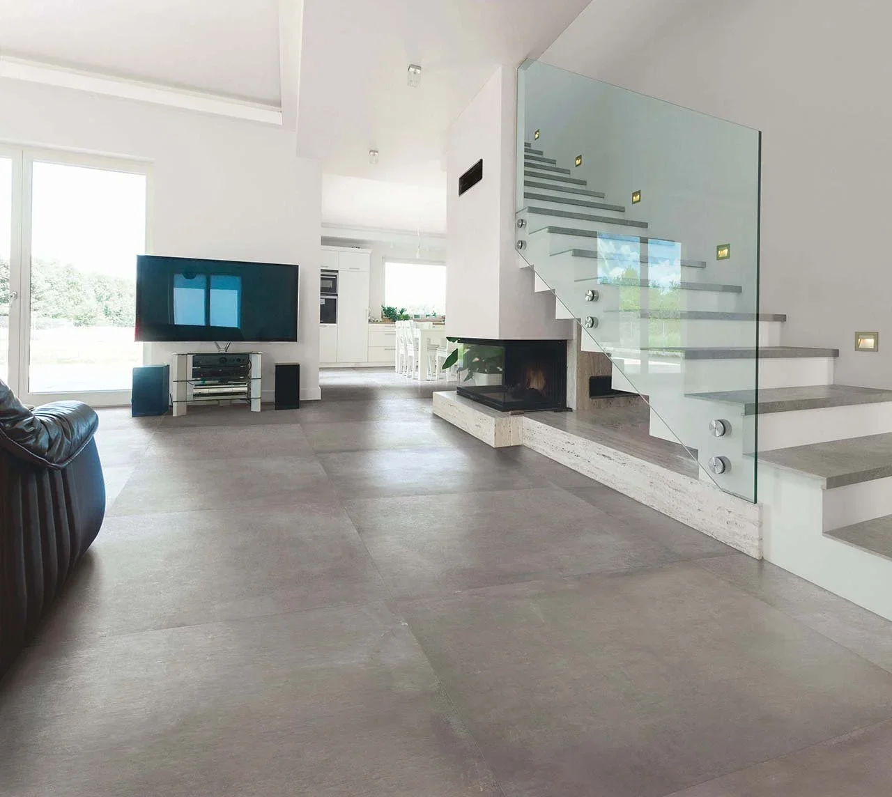 Moderne, offene Wohnzimmer mit Fernseher, Cheminée, Treppe aus Glas und Holz, große Fenster mit Blick ins Grüne, minimalistische Einrichtung.