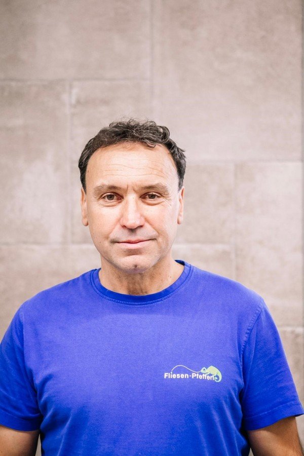 Ein Mann trägt ein blauen T-Shirt mit einem Logo und Text, die auf einen Fliesen-Fachhandel hinweisen, vor einer hellen Steinwand.