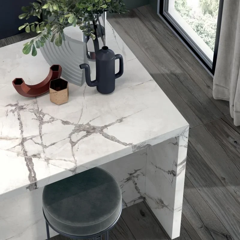 Innenraum mit Marmor-Tisch, eine Stehleiter, eine Topfpflanze, eine schwarze Wasserkanne, eine kleine Dose und eine geometrische Vase, neben einem großen Fenster mit grauer Gardine.
