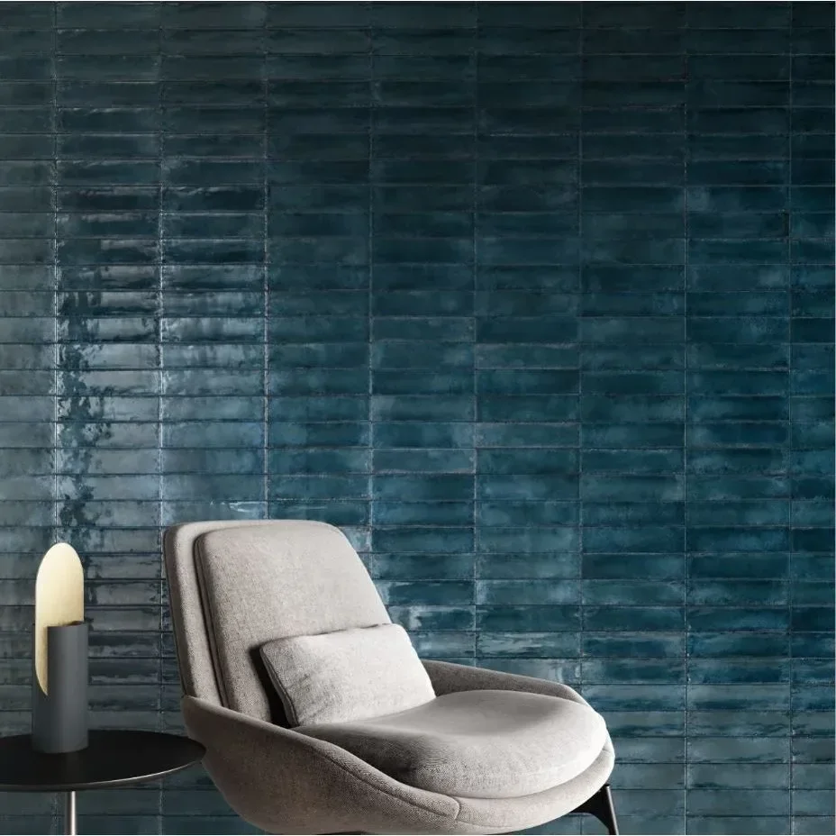 Ein graues, bequemes Sessel mit einer kleinen weichen Kissen davor, neben einem schwarzen Tisch mit einer modernen Lampe, vor einer dunkelblauen, gemusterten Wand.