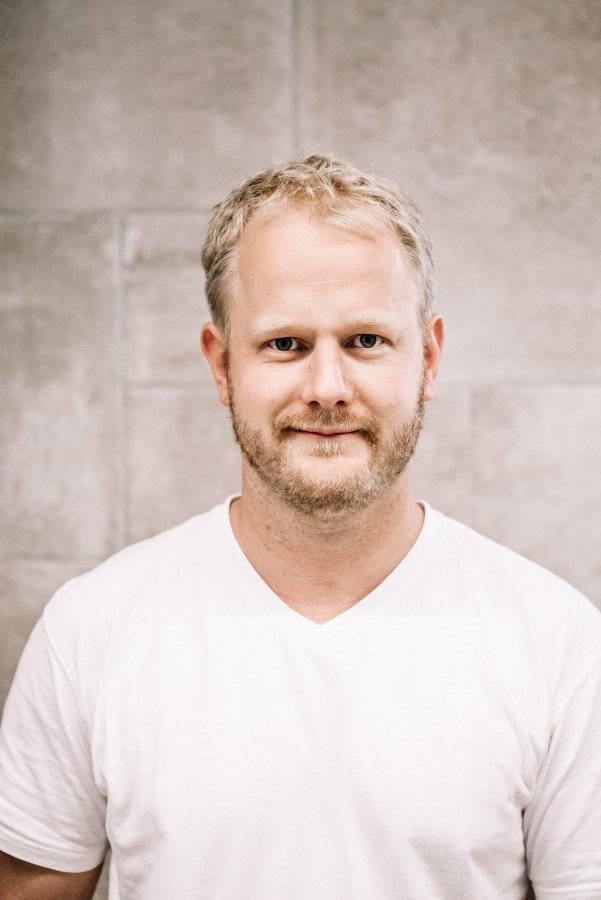 Ein Mann mit blondem Haar und Bart, trägt ein weißes T-Shirt, steht vor einer grauen Wand.