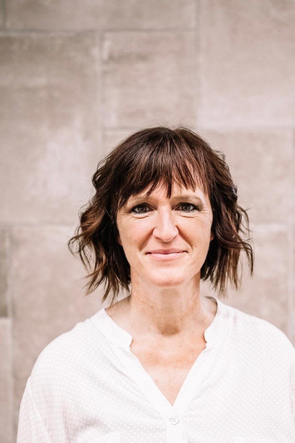 Porträt einer Frau mit braunen, kurzen, gewellten Haaren, die ein weißes Hemd trägt, vor einer hellen Wand.