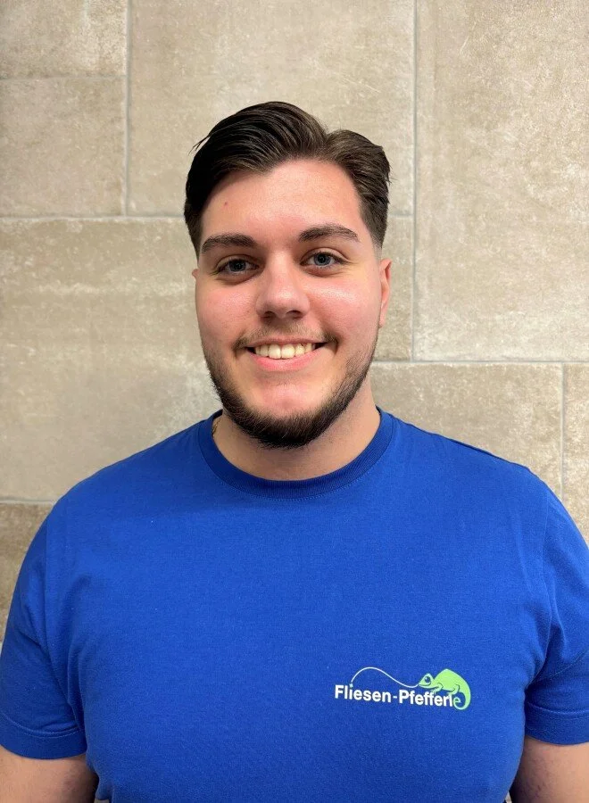 Ein lachender junger Mann mit dunklem Haar, Bart, trägt ein blaues T-Shirt mit dem Logo 'Fliesen-Pfeffer' vor einer Steinmauer.