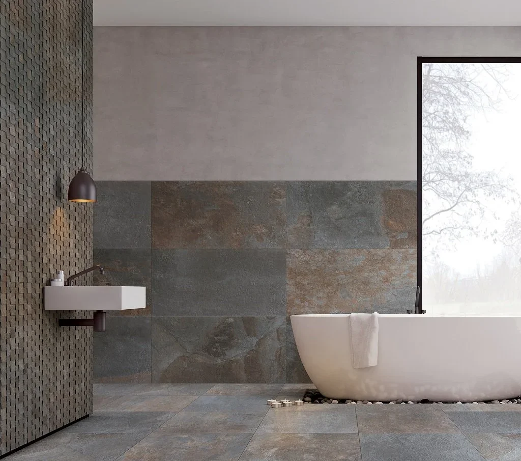 Badezimmer mit einer freistehenden Badewanne vor einem großen Fenster, eine Wandfliese mit einer Sandsteinoptik, eine kleine weiße Waschbecken-Säule mit Wasserhahn und Handseife, eine patternierte Wand und ein einfarbiger Oberputz an der Decke.