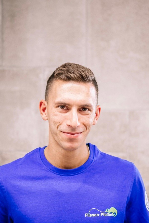 Ein junger Mann trägt ein blaues T-Shirt mit dem Logo 'Fliesen-Pfeiffer' und lächelt vor einer hellen Wand.
