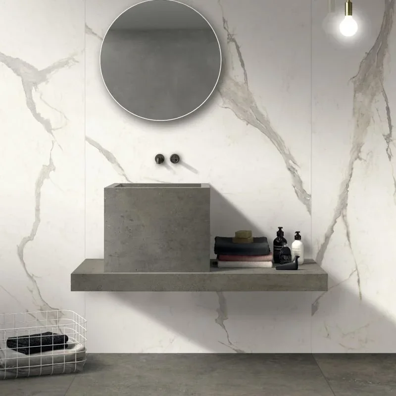 Moderner Badezimmerbereich mit einer runden Spiegel, Waschbecken auf einer Betonplatte, dunkle Seifenspender und Handtücher. Weißmarmor-Wand mit grauen Adern, einfaches minimalistisches Design.