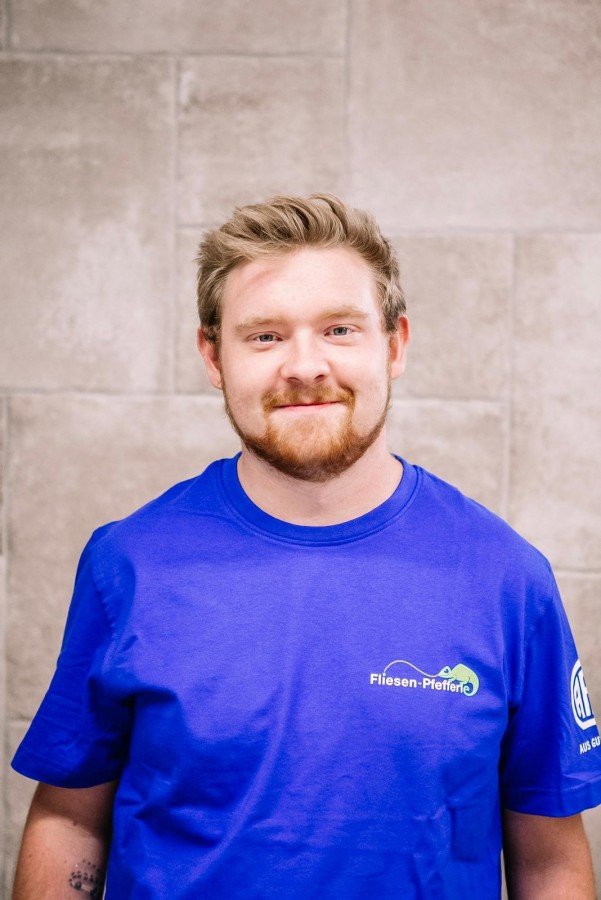 Ein junger Mann mit blondem Haar und Bart trägt ein blaues T-Shirt mit einem Logo und steht vor einer Steinmauer.