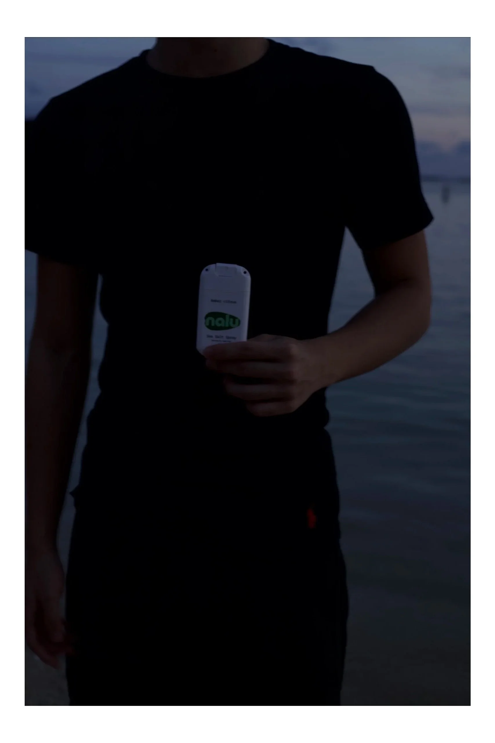 Person in a black t-shirt holding a small device near water, with a blurry background of water and sky.