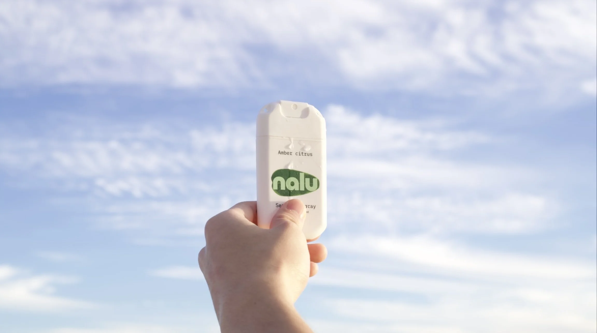 A hand holding a Nalu dry shampoo bottle labeled 'Amber Citrus' against a background of a blue sky with scattered clouds.