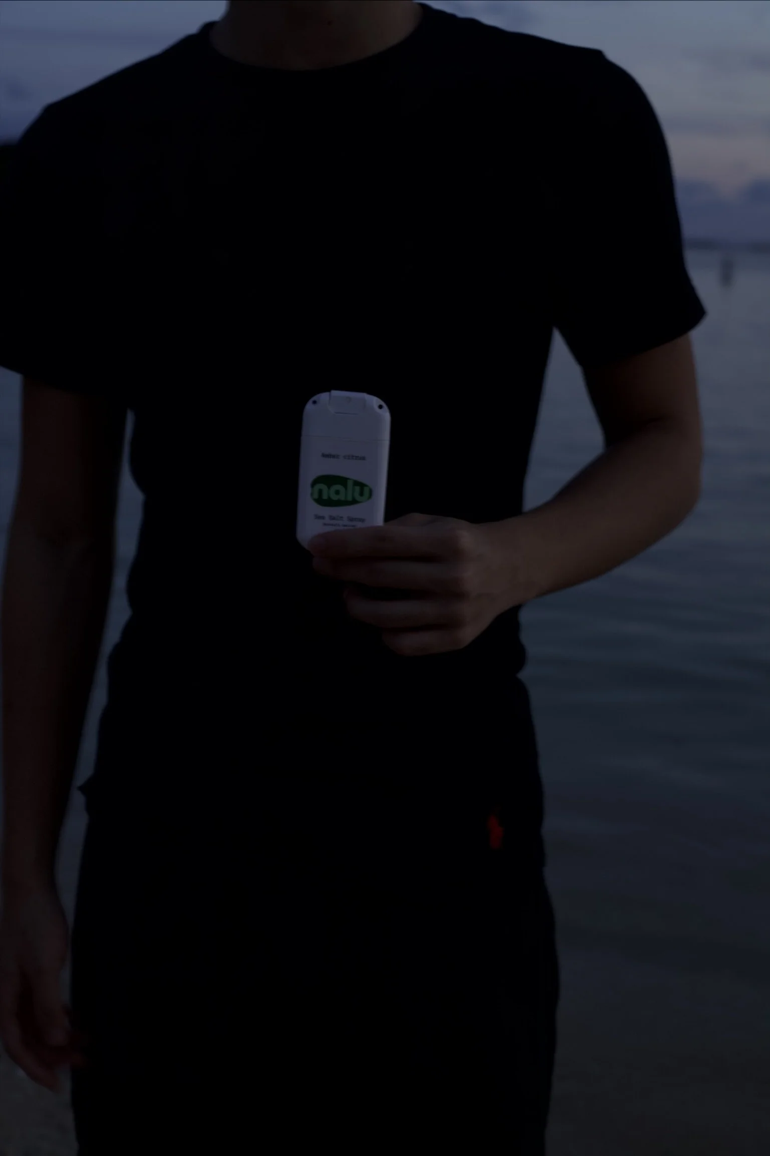 Person wearing a black t-shirt holding a small white device with a green label near a body of water during dusk.