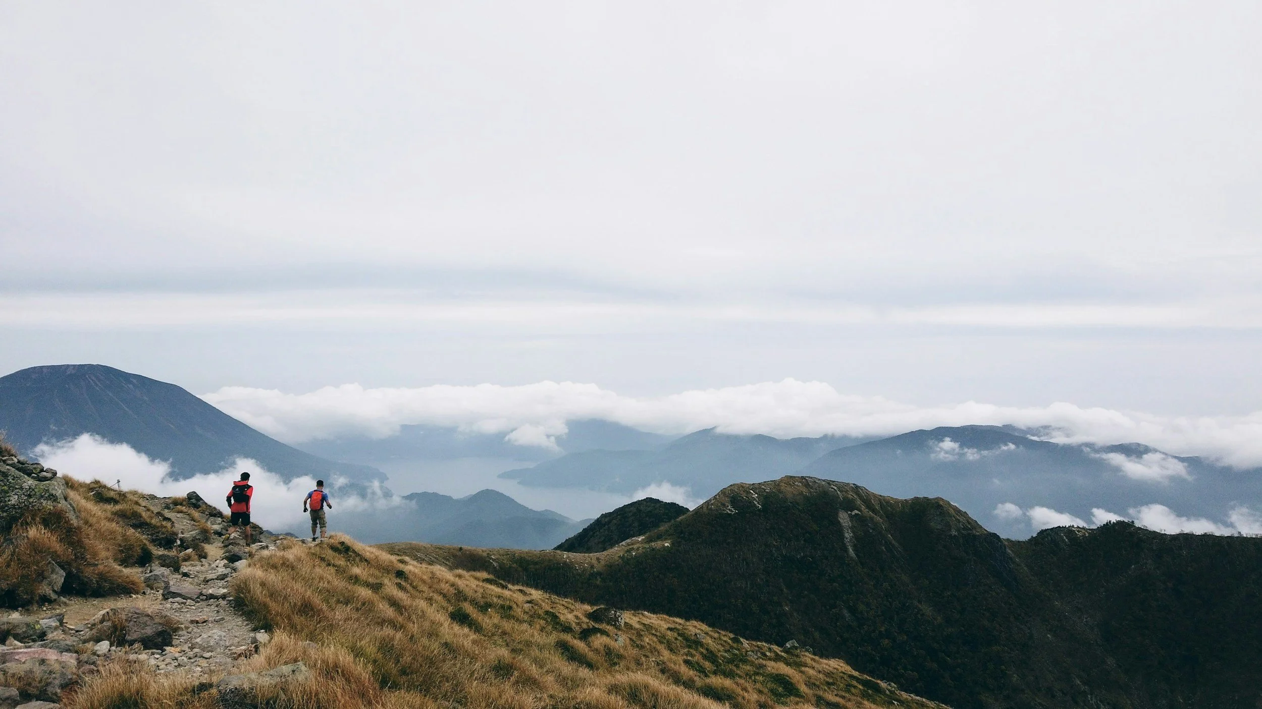 Two hikers with backpacks walking on a mountain trail through grassy terrain with cloud-covered mountains and a lake in the background.