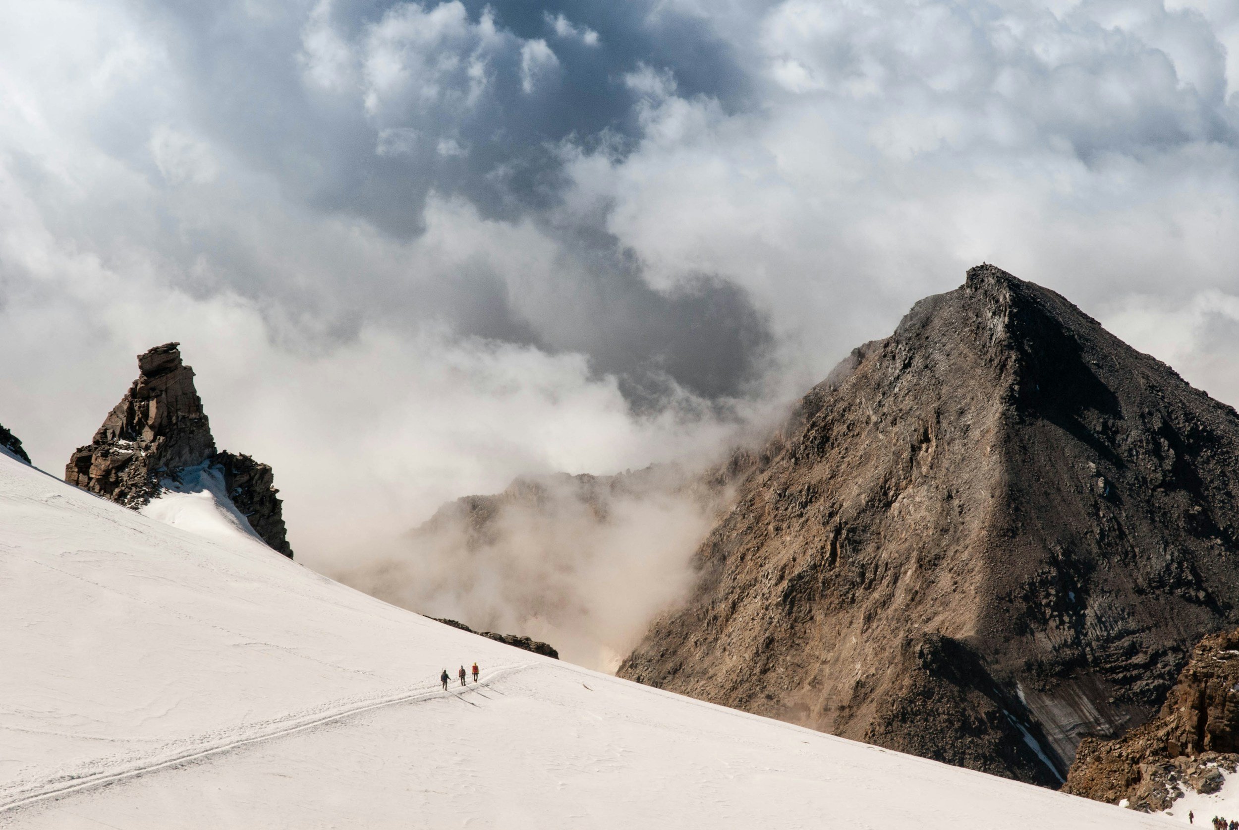 Snow-covered mountain landscape with three small hikers walking in the distance, rugged rocky peaks, and dramatic cloudy sky.