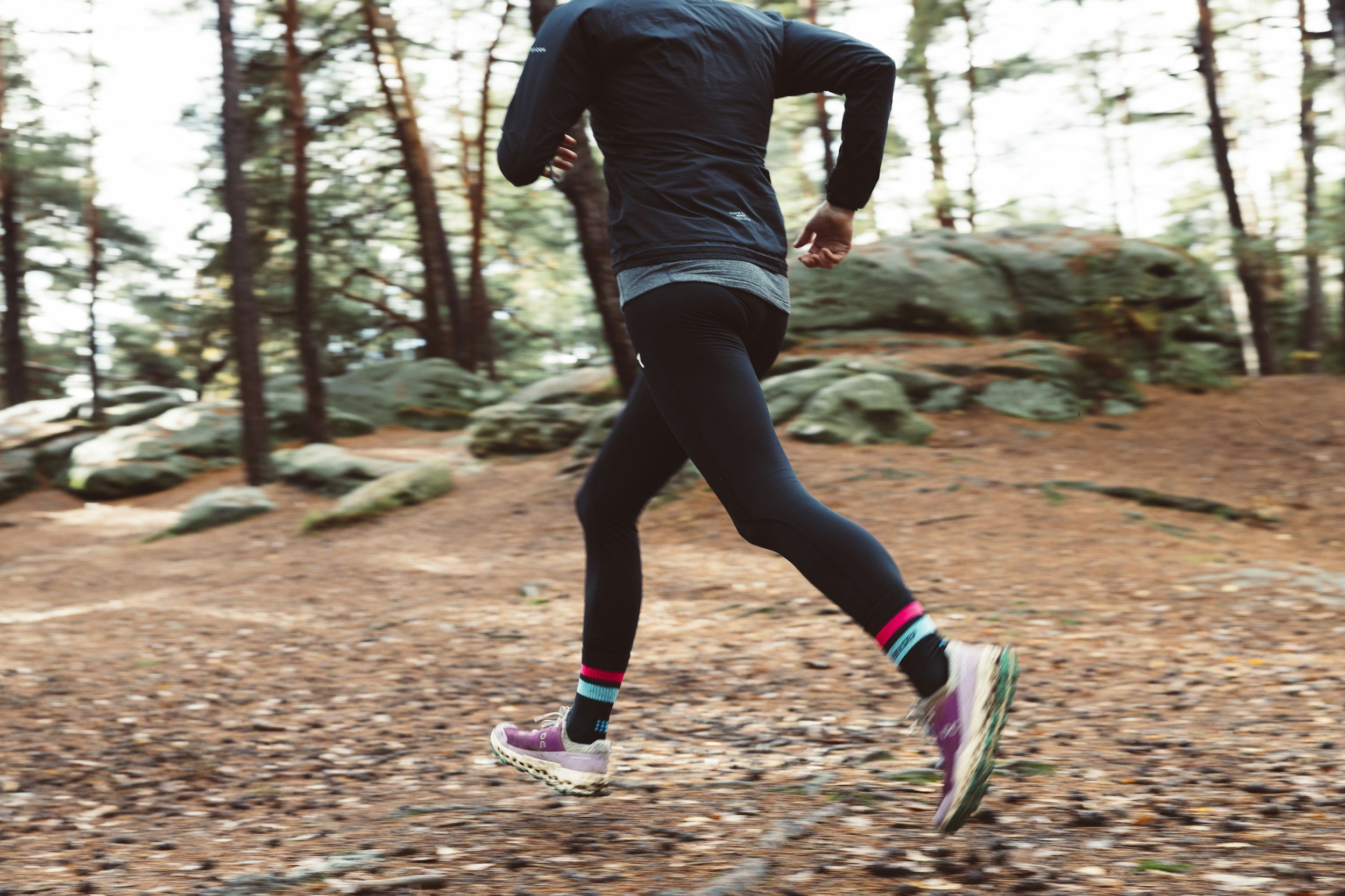 A person running on a dirt trail in a forest with trees and rocks in the background.