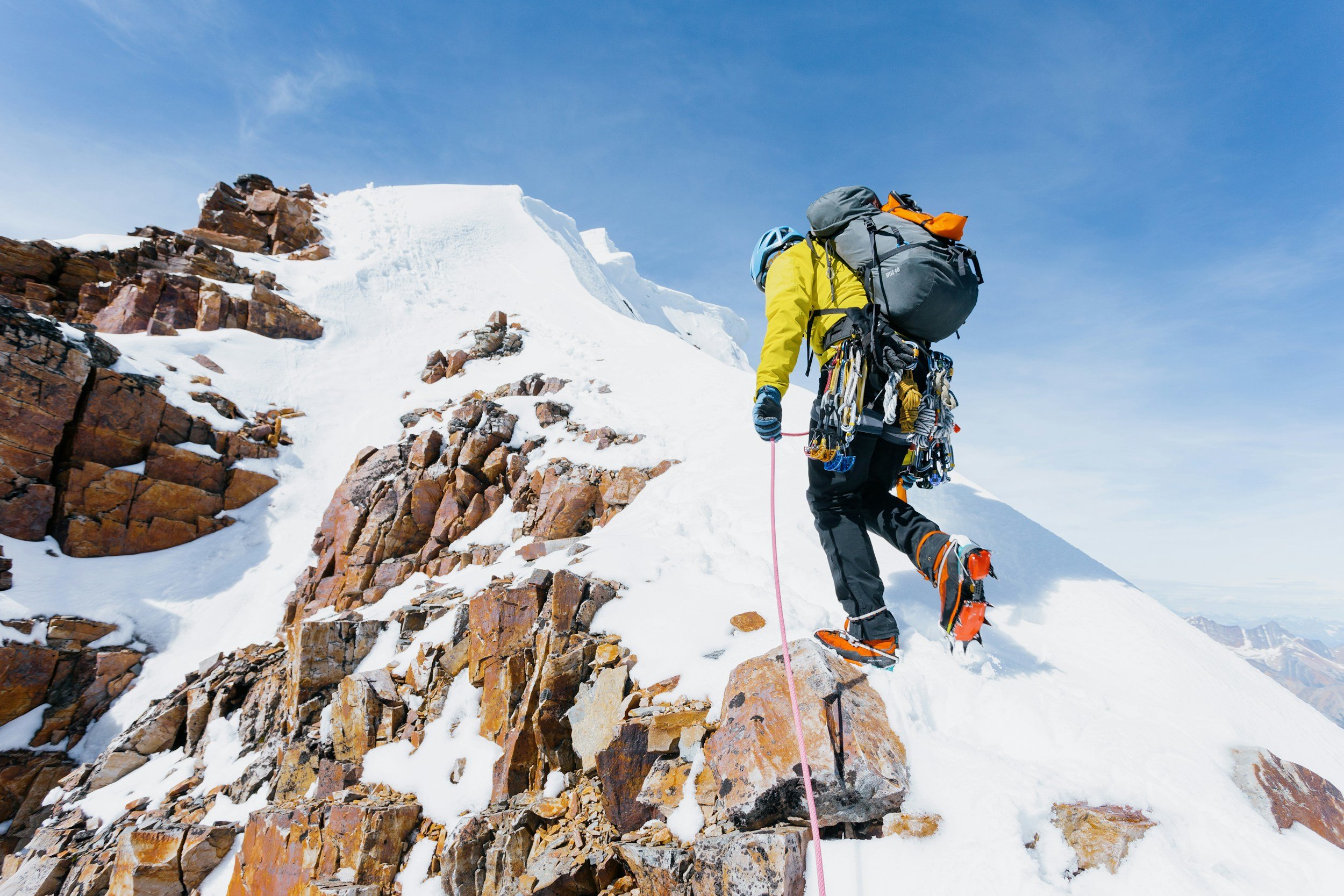 A climber ascending a snow-covered mountain ridge with climbing gear, backpack, and safety equipment, under a blue sky.