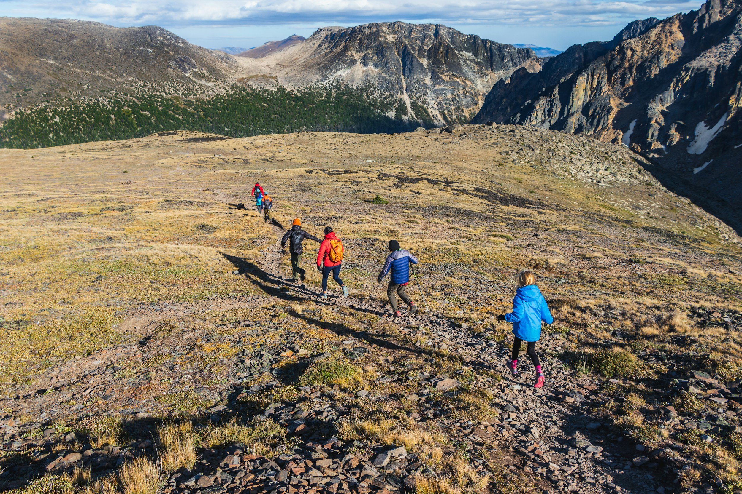 A group of seven hikers trekking on a mountain trail in a rugged landscape with rocky terrain and mountains in the background.