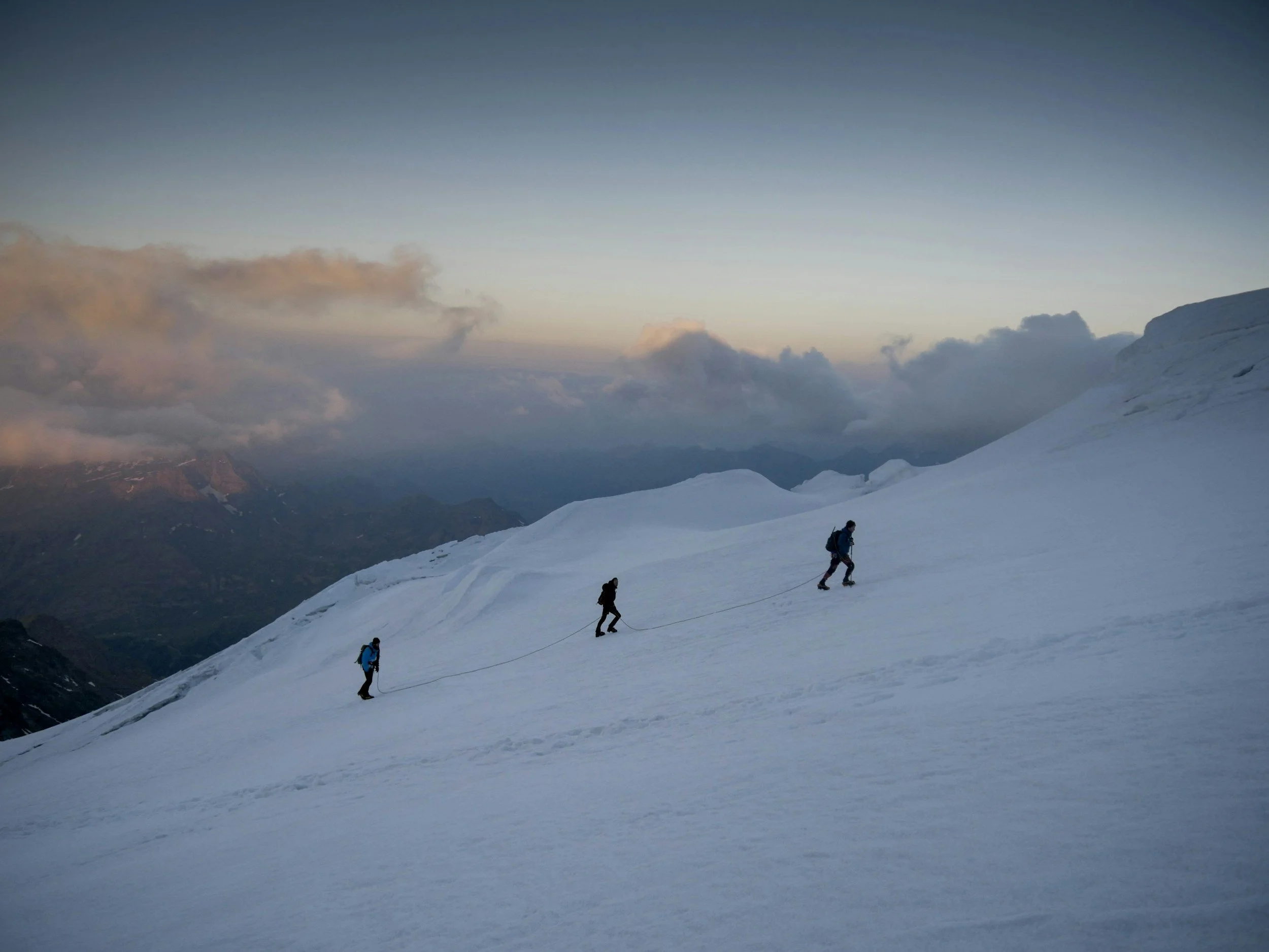 Three climbers ascending a snow-covered mountain slope during dusk or dawn with a cloudy sky and distant mountains.
