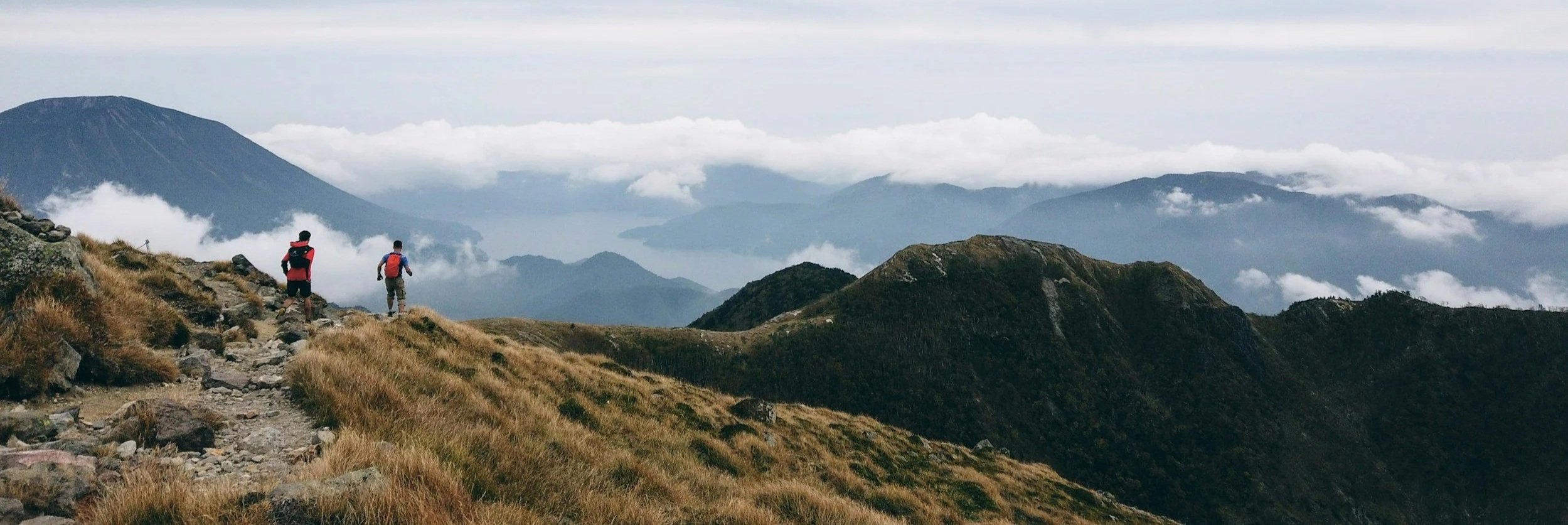 Two hikers walking on a rocky mountain trail with grassy slopes, overlooking distant mountains and clouds.