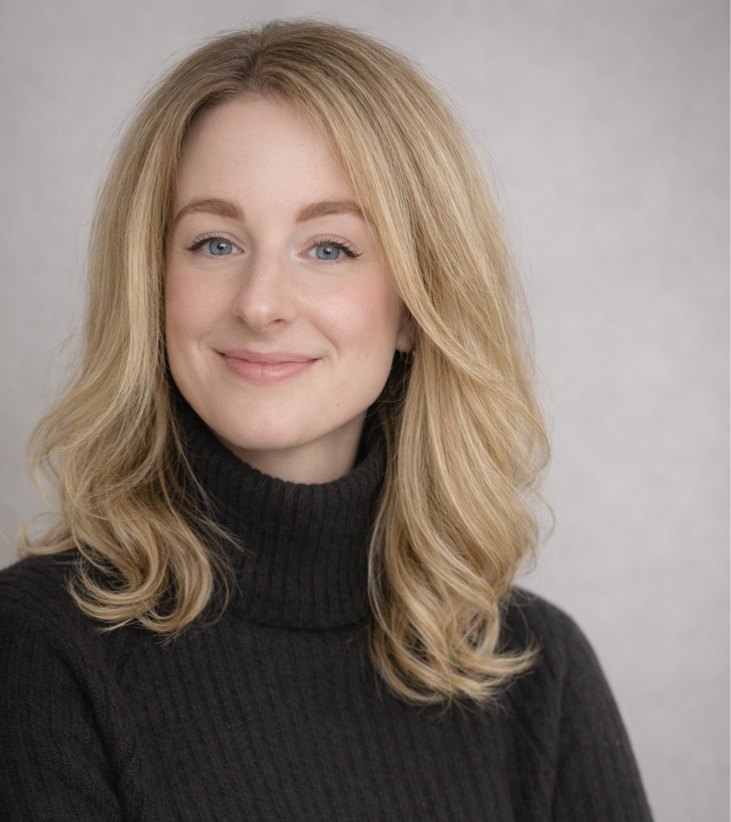 Nahaufnahme eines lachenden jungen Frau mit blonden, gewellten Haaren, hellen Augen, in einem schwarzen Rollkragenpullover vor einem neutralen grauen Hintergrund.