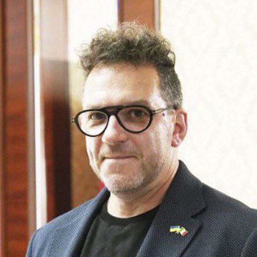 A man with curly hair wearing glasses, a black shirt, and a blue blazer with a small flag pin, standing indoors against a blurred background.