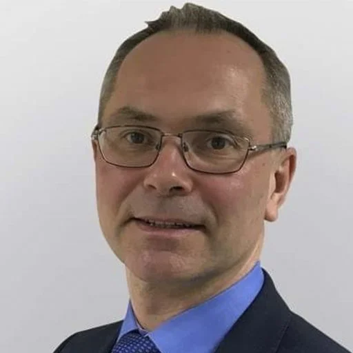 A headshot of a man with short hair, glasses, wearing a suit and blue shirt, against a plain light background.