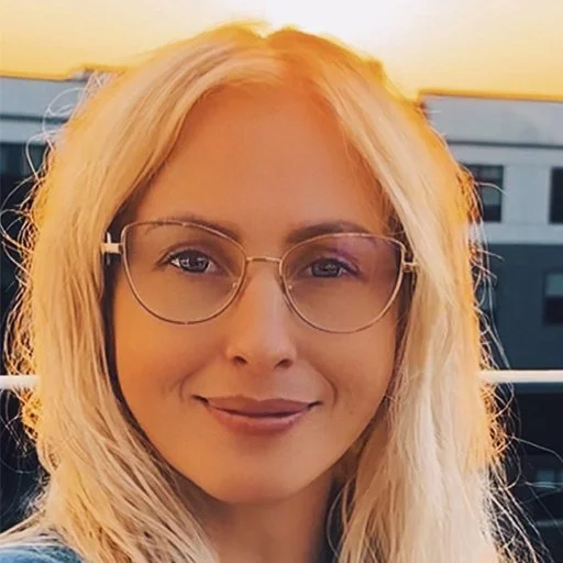 Close-up of a woman with blonde hair, wearing glasses and smiling, outdoor setting with a railing in the background.