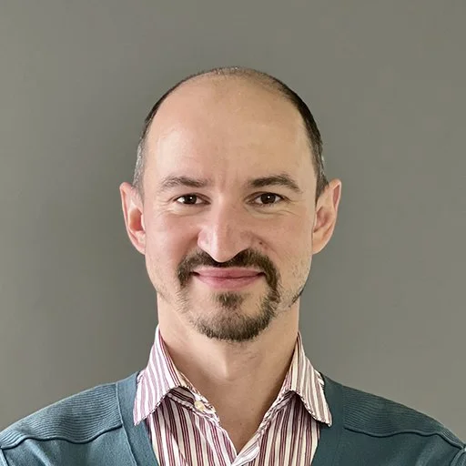 Portrait of a man with a beard and mustache smiling, wearing a striped shirt and a blue sweater, against a plain gray background.