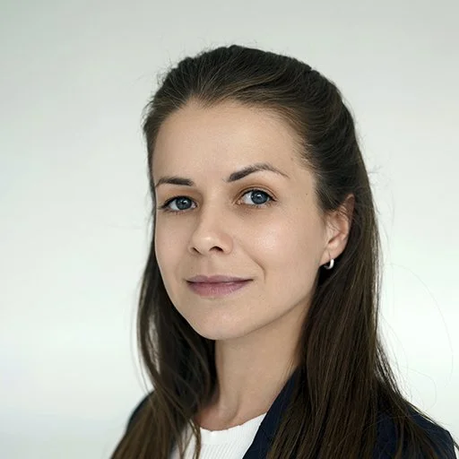 A young woman with long brown hair, blue eyes, and fair skin, wearing a black blazer and small earrings, smiling softly against a plain white background.