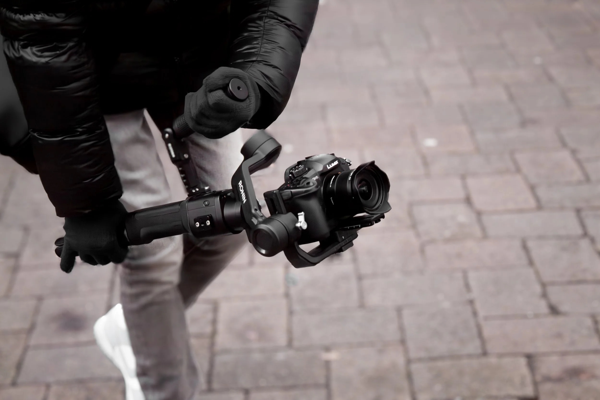 Person holding a camera stabilizer with a camera attached, walking on a brick sidewalk.