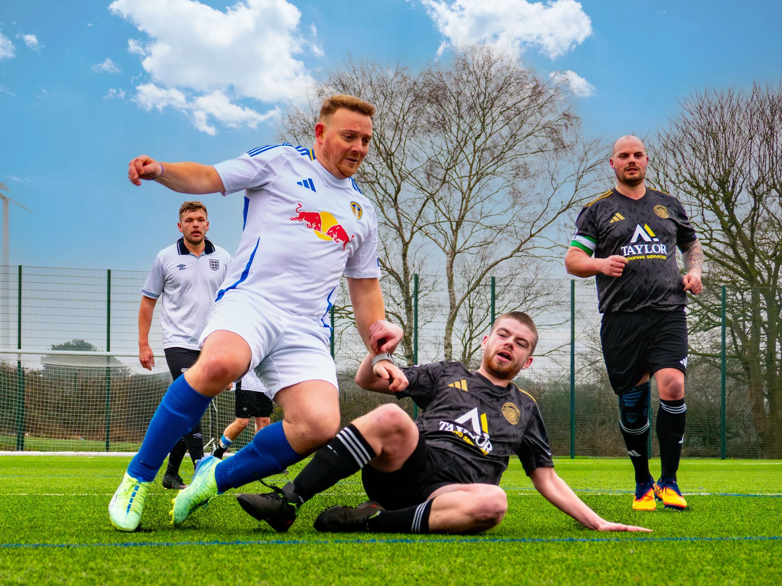 Football players on the field, one player in a white football shirt falling over another player in a black football shirt who is on the ground, with two additional players in black and white football shirts running.