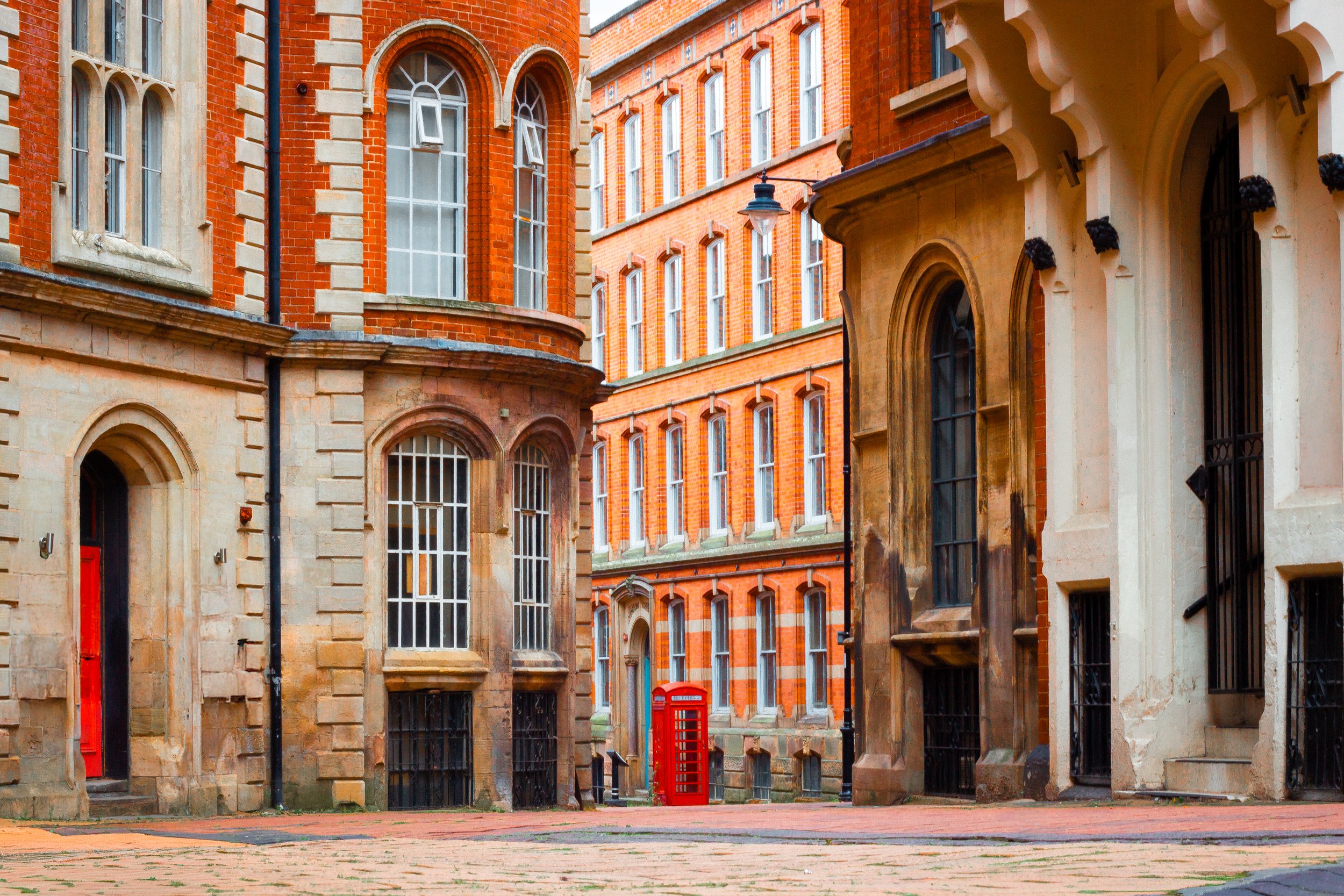 A narrow alley with historic red-brick buildings and large arched windows, featuring a red telephone booth in the distance.