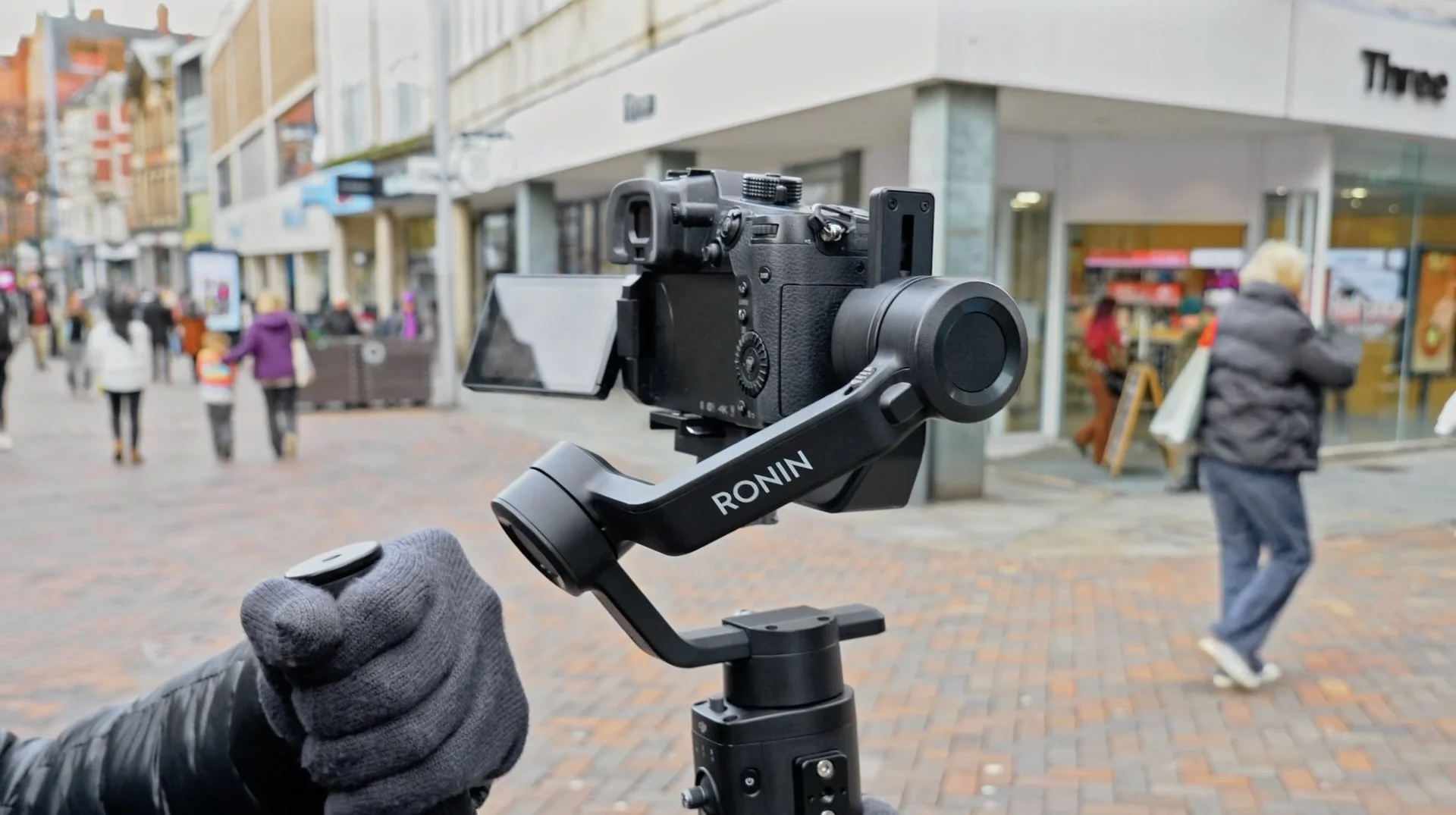 A camera mounted on a Gimbal stabilizer with a person's hand controlling it, set on a busy pedestrian street with storefronts and people walking in the background.