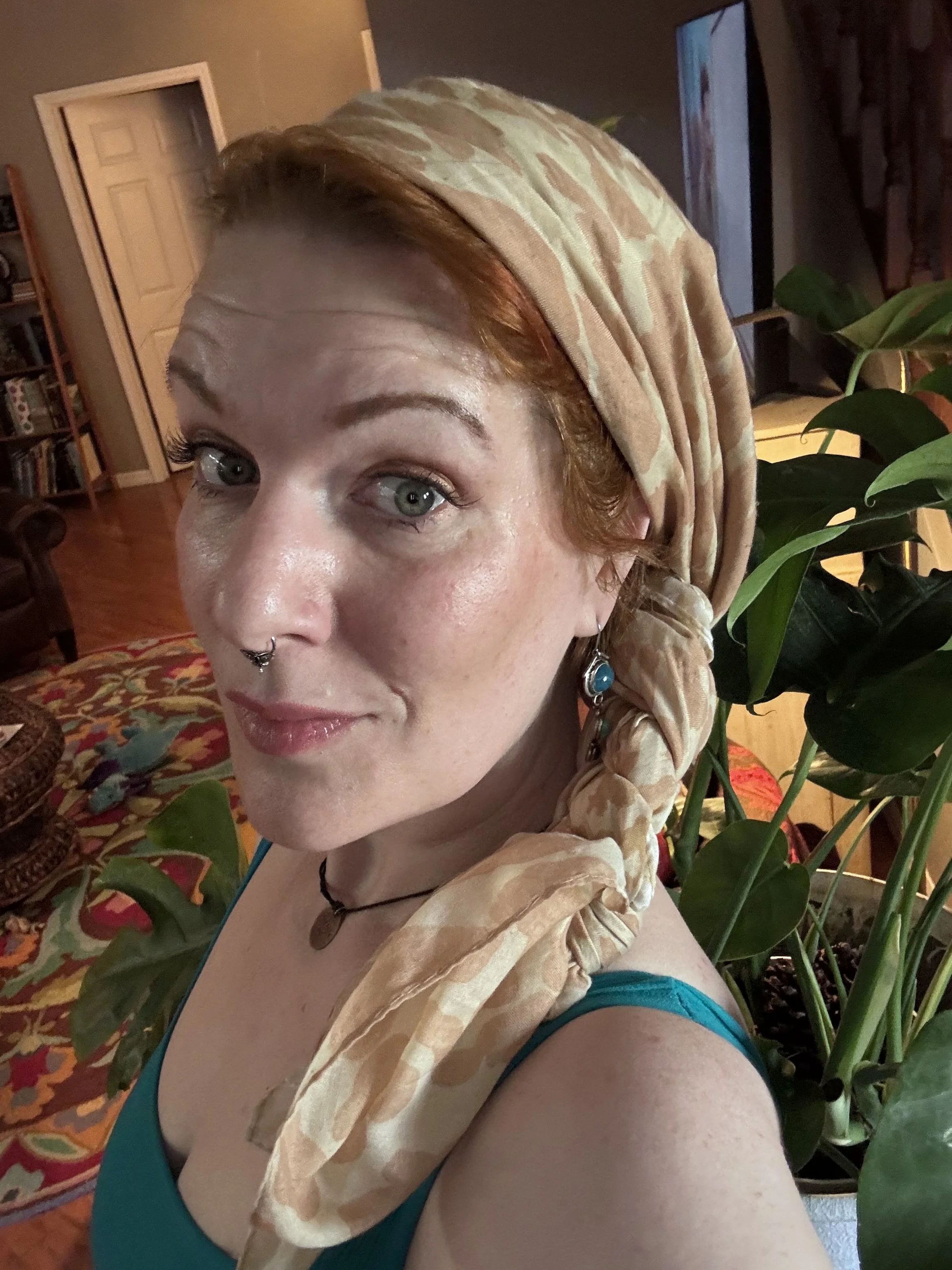 Ivy Godiva in a relaxed, candid selfie — wearing a sandy-toned head wrap and teal top, with lush houseplants visible behind her, a nose ring, and a soft, open expression