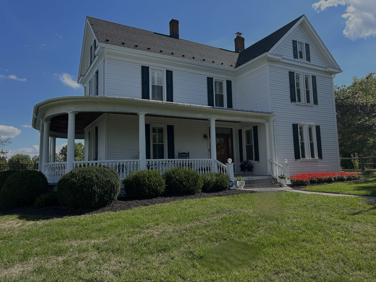 Completed Exterior Painting Job by CMT Painting Co. A large, white, two-story house with blue shutters, a wrap-around porch with columns, and a well-maintained lawn with bushes and flowers, under a partly cloudy sky.