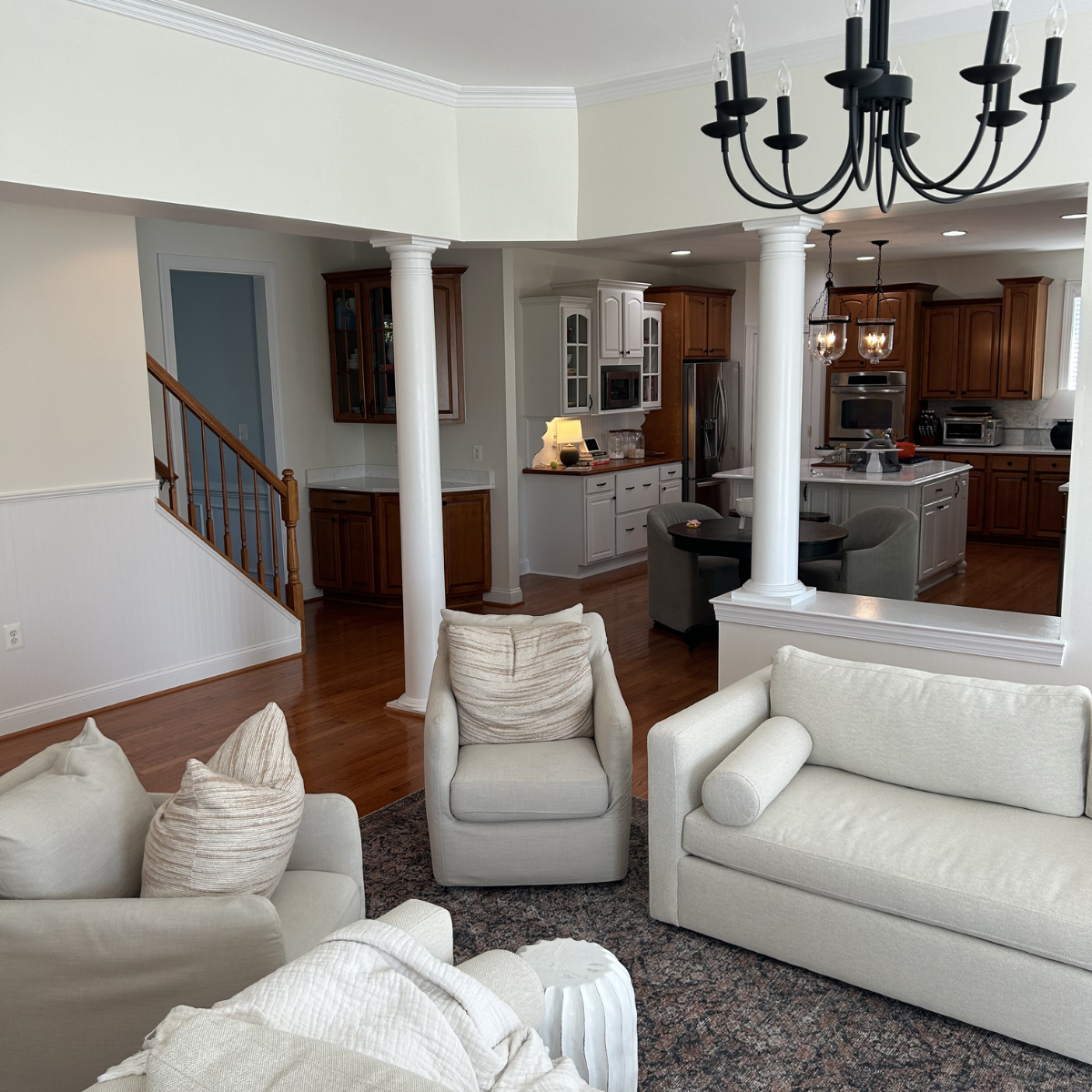 Spacious living room with white upholstered furniture, a dark wooden dining table with chairs, and an open kitchen with wooden cabinets and stainless steel appliances. Two white columns separate the living room from the kitchen. Chandeliers hang from