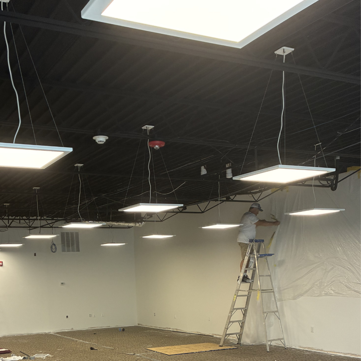 CMT Painting Commercial Painting Winchester.  A worker standing on a ladder installs or repairs ceiling lights in a room, with a white wall and wooden ceiling, and some construction materials on the floor.