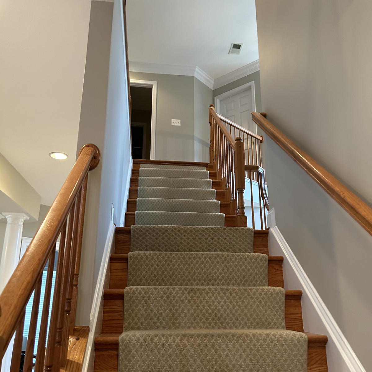 CMT painting Co staircase painting in Winchester, VA. Project: Interior view of a staircase with wooden railing, carpeted steps, and a light gray wall with a door at the top.