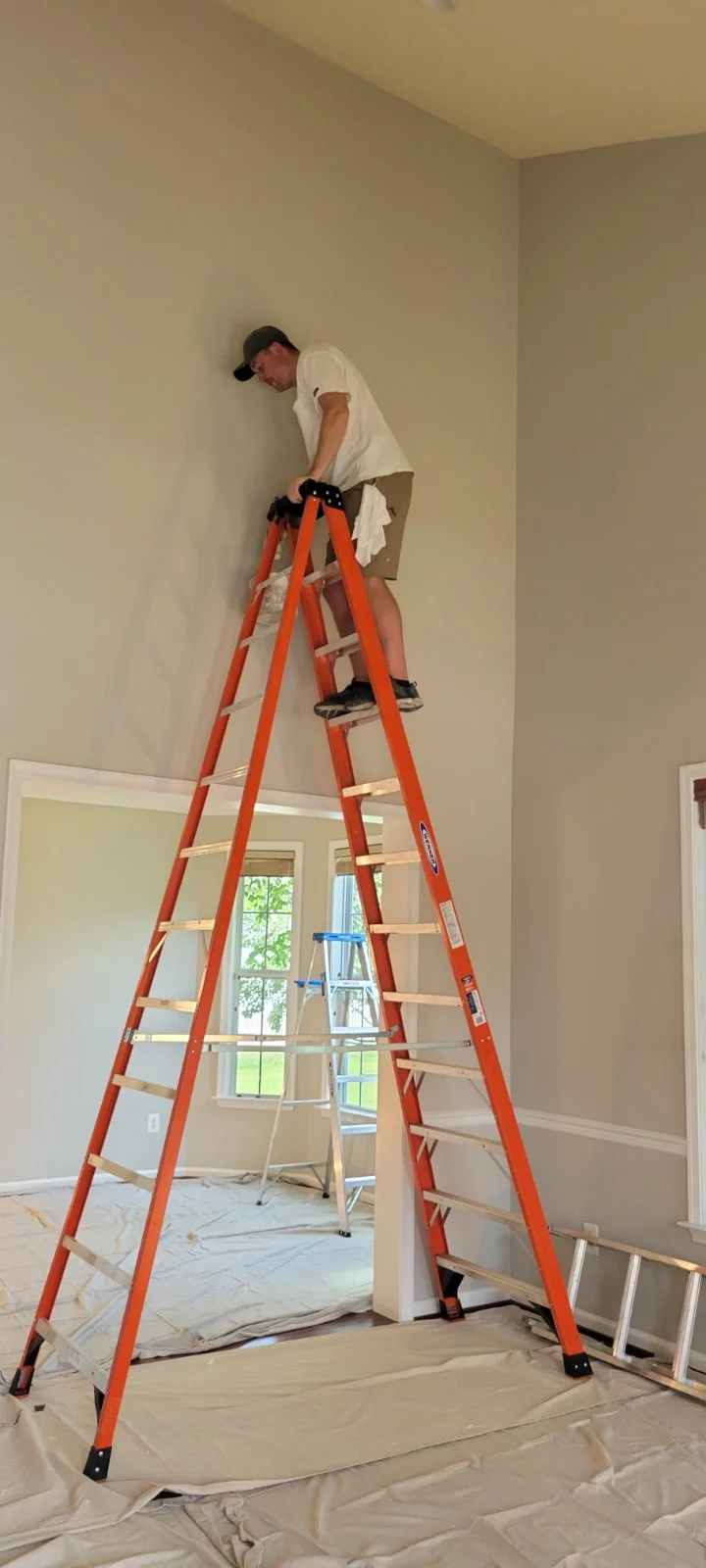 Winchester VA interior painting CMT Painting Co. A man standing on an orange ladder, painting or working on the wall near the ceiling in a room with beige walls and large windows, with another smaller ladder and drop cloths on the floor.