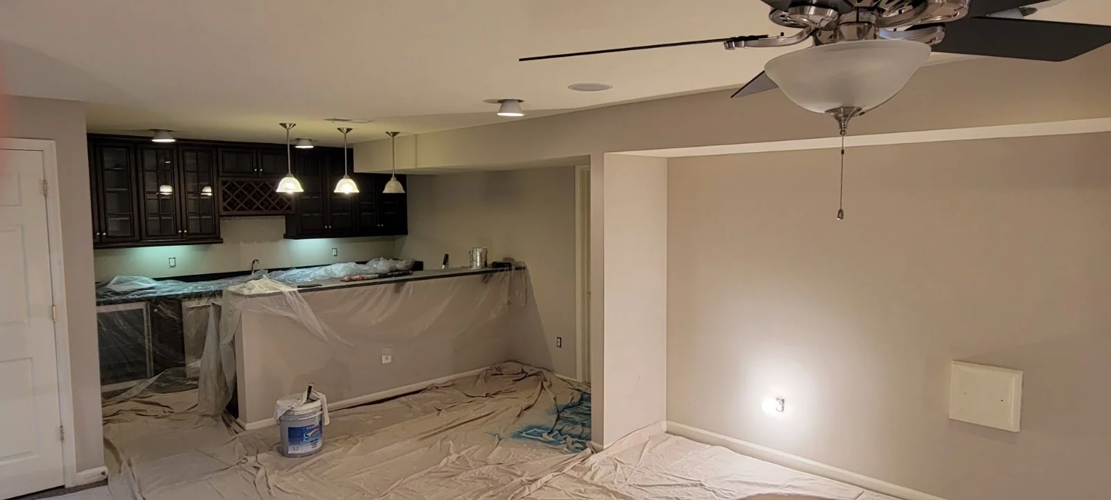 Interior painting Winchester Virginia A room under renovation with plastic sheets and painter's tape on the floor and cabinets. There are hanging lights over a counter area, and a ceiling fan is visible.