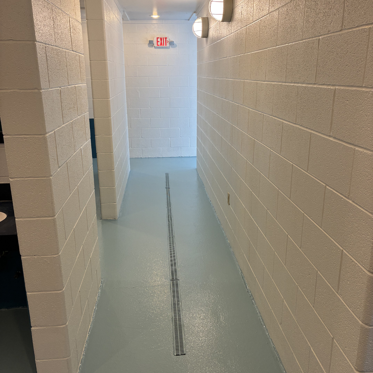 Empty hallway with beige cinder block walls, a light blue floor, a drain in the center, two wall-mounted lights, and an exit sign at the end. CMT Painting Co. commercial painters Winchester VA 