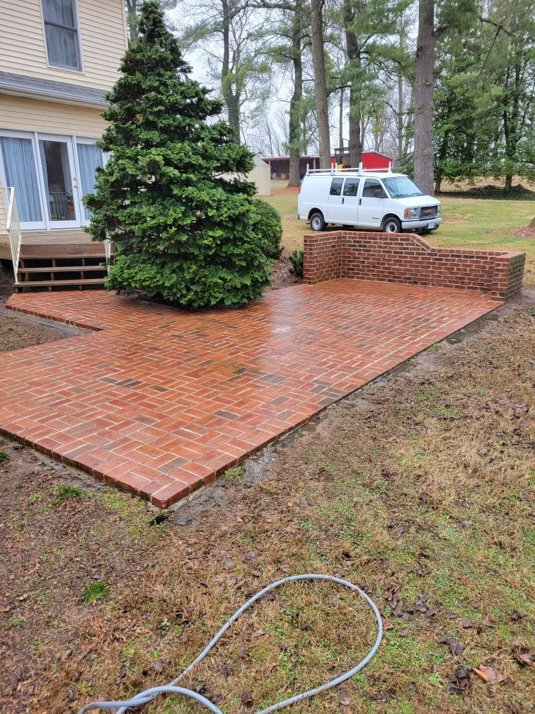 Pressure washing Winchester VA. A newly poured brick patio next to a house with a large evergreen tree and a white van parked nearby. The patio has a small brick half-wall on one side, and a garden hose is on the ground in the foreground.