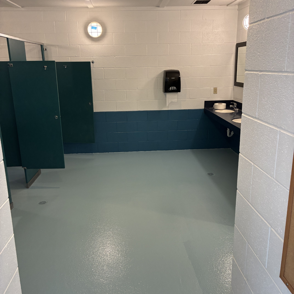 Commercial painters Winchester Virginia – Restroom with blue and white tiled walls, a black countertop with two sinks, a mirror, a paper towel dispenser, a bathroom stall, and a drain in the floor.
