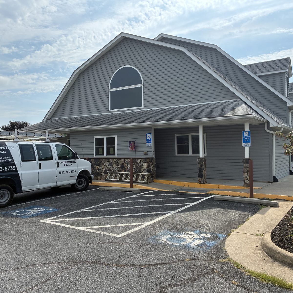 A gray building with a sloped roof and an arched window at the top. There is a parking lot in front with a parking space designated for handicapped parking, marked by blue signage and a painted symbol on the ground. A white service van is parked in o