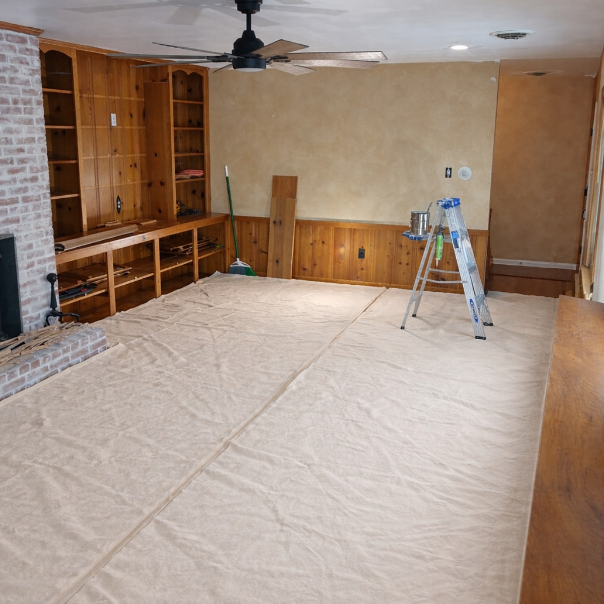 A room undergoing renovation with a ladder, paint supplies, and empty wooden bookshelves.