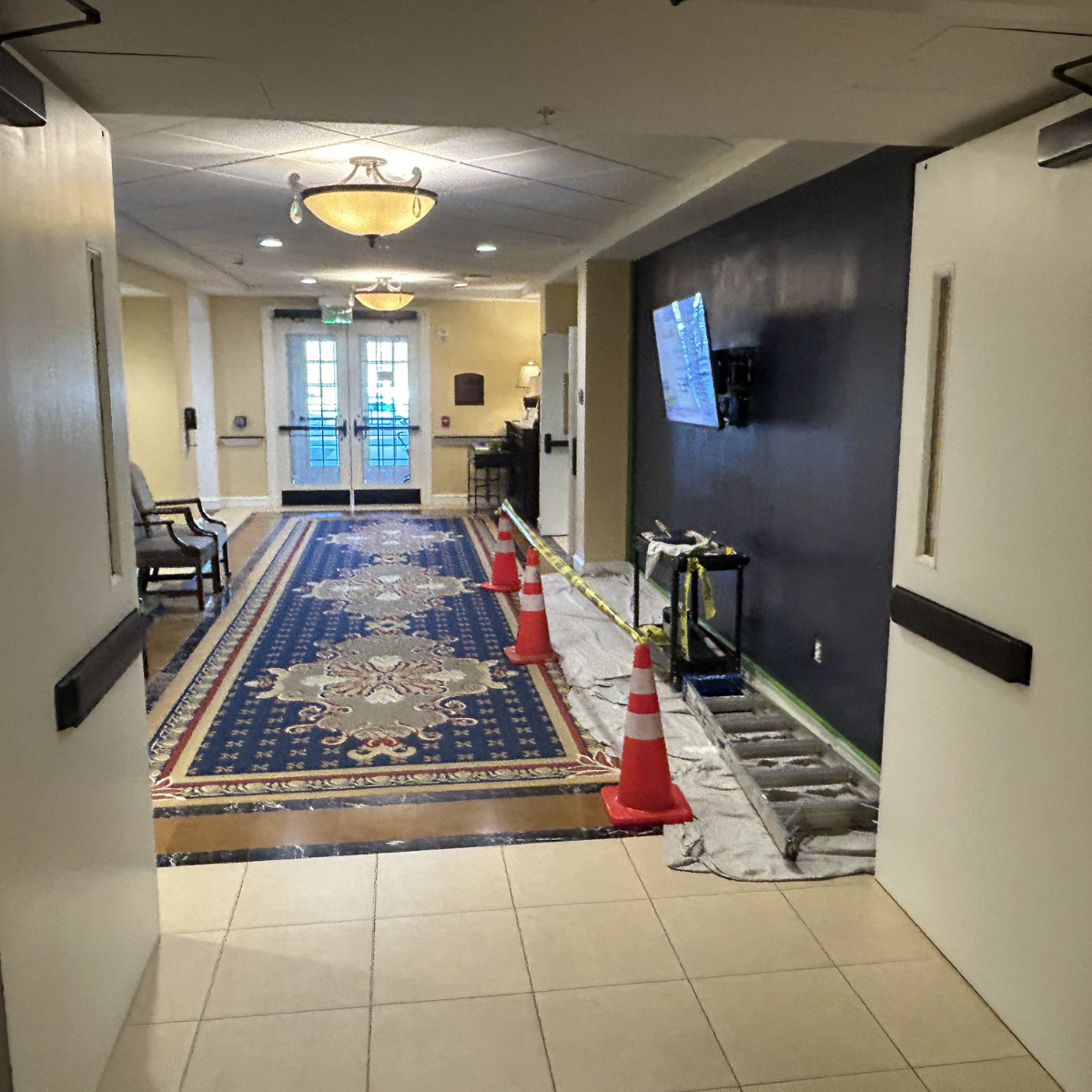 Commercial painting services Winchester Virginia - Hallway in a building with a patterned rug, chairs, and a dark accent wall with a mounted TV. Construction cones, barriers, and a ladder indicate ongoing repairs.