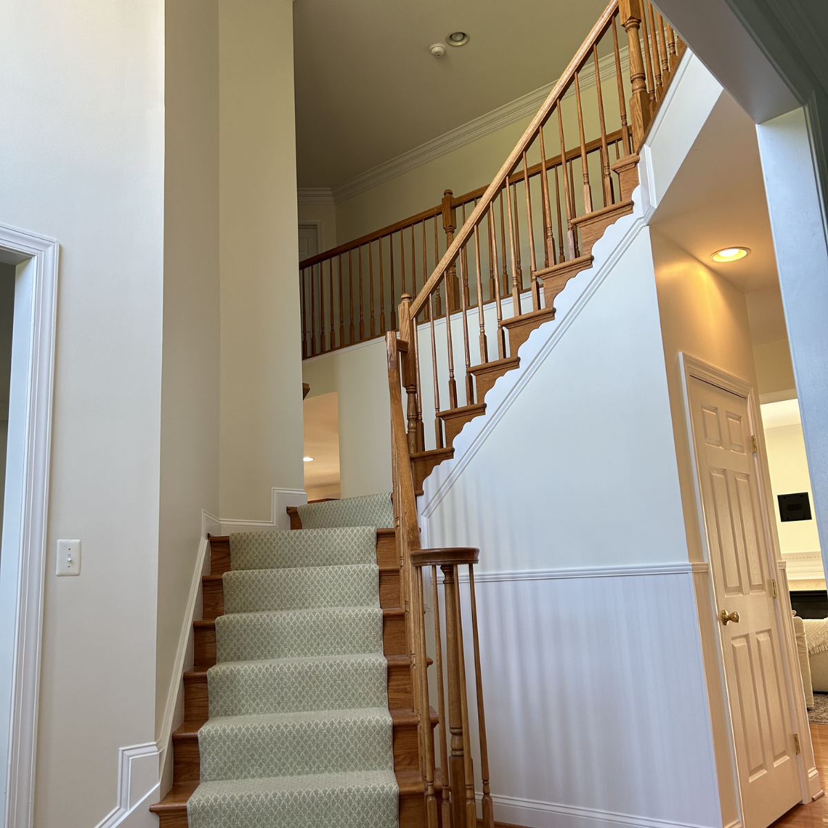 Interior view of a staircase with wooden handrails and newel posts, a beige carpet runner, and off-white walls with white trim. A door is partially visible on the right.